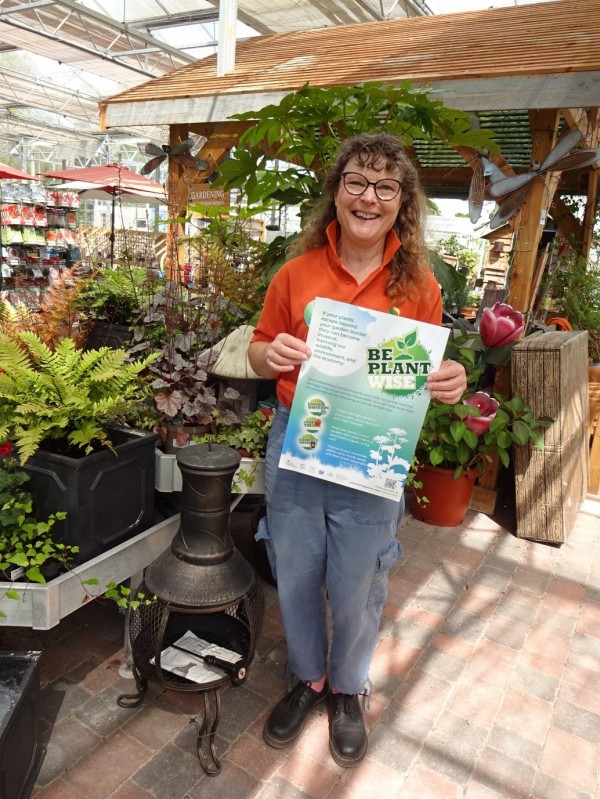 A woman with red curly hair stands in front of a group of plants in a garden centre holding a poster that reads 'Be Plant Wise'