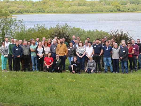 A large group of people stand on grass infront of a lake