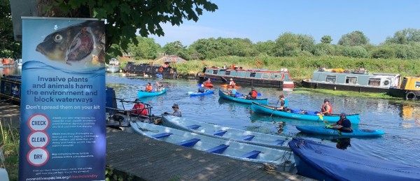 A group of kayakers are out on a canal in five blue kayaks. Some green floating plants are visible behind them. Three narrowboats are on the other side of the river and in the foreground a pop up banner reads 'Invasive plants and animals harm the environment and block waterways, don't spread them on your kit. Check, Clean Dry'.